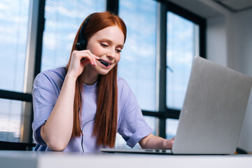 Close-up face of smiling young woman operator using headset and laptop during customer support at home office. Young female student communicating online by video call on background of window.