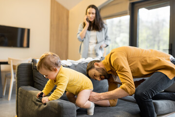 Man wearing his baby son on the couch at home. Fatherhood and child care by father concept. Wife talks on phone on the background