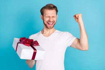 Portrait of astonished cheerful person fist up closed eyes hold giftbox isolated on blue color...