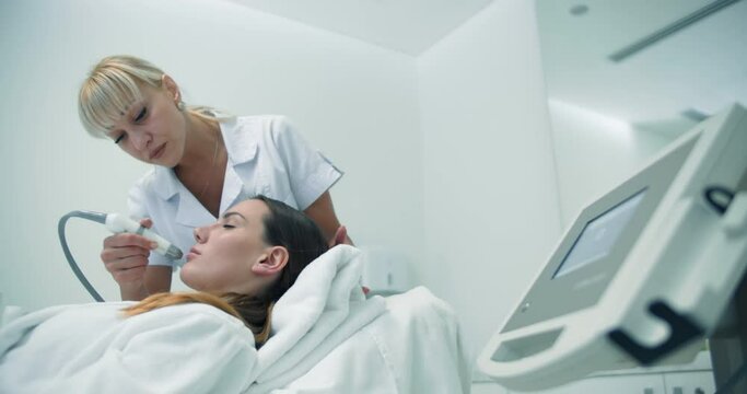 A female cosmetologist is using the Dermadrop TDA device for doing a non-injection bio-revitalization procedure on the face of a female client who is laying in the cosmetology bed.
