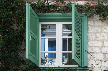 A blue wooden traditional window is reflected in the glass of a green wooden window, on a stone wall full of climbing plants. Nafplio, Greece