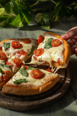 hand raising a slice of homemade pizza with cherry tomatoes and fresh basil on wooden board and plants in the background with sunlight