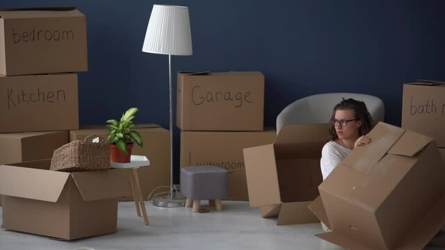 Portrait of an adult woman lying on the floor under empty cardboard boxes. Moving difficulties, decluttering and minimalism concept. Funny video
