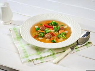 Homemade lunch. Vegetable soup with meatballs, broccoli, carrots, celery, potatoes, red pepper and herbs in a white plate on a white wooden table, napkin and spoon. Background image, copy space