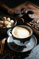 Morning coffee. Cappuccino in a small cup with a spoon on a saucer. Sugar, cookies, chocolate, cinnamon, coffee beans on a wooden board on a black table. Sunlight. Background image, copy space
