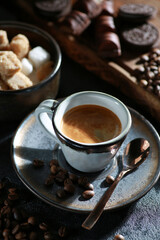 Morning coffee. Espresso in a small cup with a spoon on a saucer. Sugar, cookies, chocolate, coffee beans on a wooden board on a black table. Sunlight. Background image, copy space