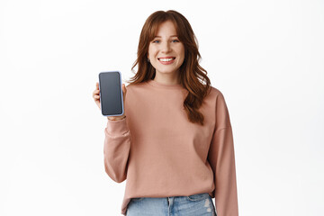 Fototapeta premium Portrait of happy cute girl laughing, showing phone screen and smiling, recommend app for smartphone, standing against white background