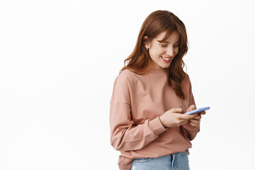 Naklejka premium Smiling natural ginger girl, chatting with smartphone, looking at phone and messaging, standing in casual clothes against white background