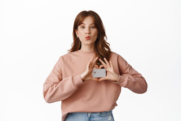Silly redhead girl pucker lips kissing, showing heart sign around credit card, shopping, paying for purchase contactless, standing against white background