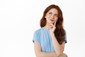 Thoughtful young woman with red hair thinking, looking up and smiling while having interesting idea, planning something, making decision, pondering while standing against white background