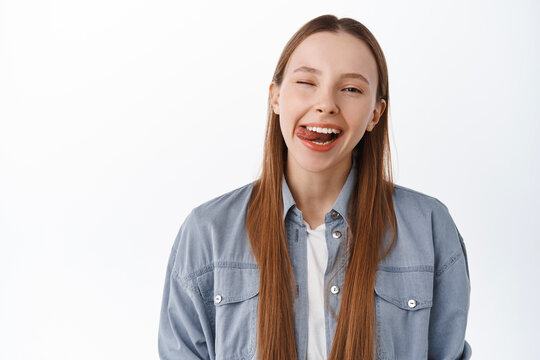 Happy beautiful teeager, girl showing tongue, smiling and winking at camera carefree, positive and joyful woman, standing over white background