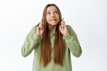 Excited cute girl biting lip nervous, cross fingers and look up at copyspace, waiting for results with worried expression, anticipating news, praying, standing over white background