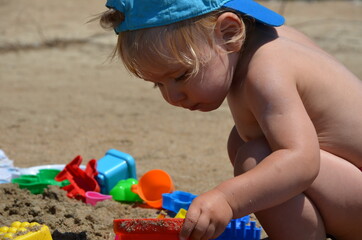 Little blond toddler boy on the beach play with sand and toys near the sea. Copy space. Summertime. Close up. Sea view. Sunshine. 