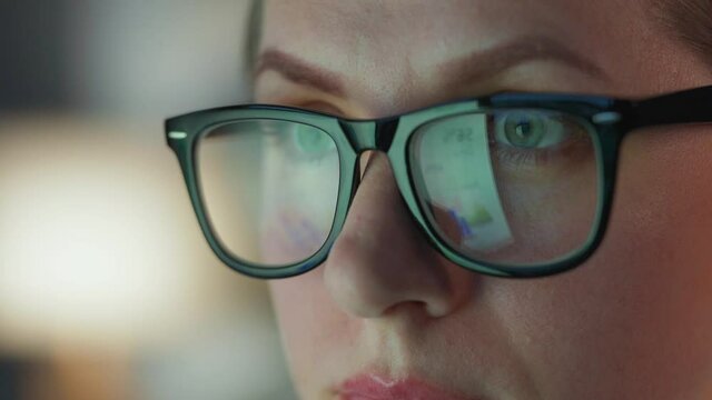 Woman in glasses looking on the monitor and and working with charts and analytics. The monitor screen is reflected in the glasses. Work at night. Extreme close-up