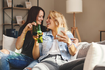 Two young happy women sitting on the sofa drinking beer and taking selfies