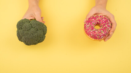 Comparison of eating habits. Woman holding broccoli and donut on a yellow background. Cope space.