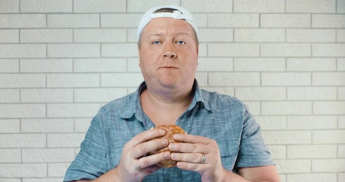 A man in a shirt and a white baseball cap happily bites a bun with a cutlet several times, chews it and swallows it. It stands against a white brick wall.