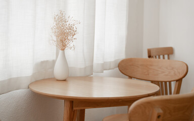 White vase with small flowers place on wooden table next to the window with white curtains and warm...