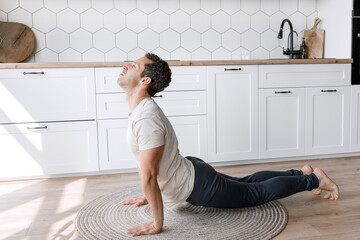 Fototapeta na wymiar Focused guy doing yoga exercises during home training. Concentrated man doing cobra pose at carpet on the floor in modern kitchen, relieve stress doing physical exercises