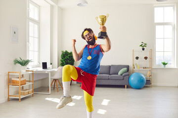Joyful emotional athlete in funny bright sportswear, who has a medal around his neck and holds a winning cup in his hand and flaunts it. Concept of victory, active lifestyle and training at home. - obrazy, fototapety, plakaty