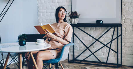 Thoughtful female in optical spectacles for provide eyes protection holding education textbook looking away and thinking in loft apartment with stylish design, young reader with literature best seller