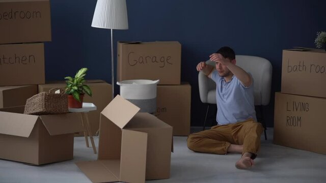 Tired Annoyed Adult Man Sits On The Floor Against The Backdrop Of Cardboard Boxes With Things. The Guy Pushes The Boxes Away From Him. Moving Difficulties, Mortgage And Debt Problems