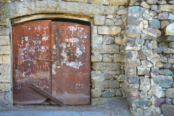 A red, old, iron, closed door with a locked bolt against the background of an old crumbling stone wall. The door has two wings. The stone wall is made of stones of different colors. Cracks are visible