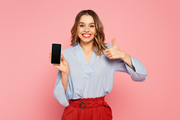 Stylish woman showing like and holding smartphone with blank screen on pink background