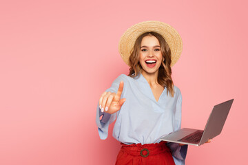Cheerful woman in straw hat pointing with finger while holding laptop isolated on pink