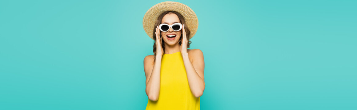 Happy woman in straw hat holding sunglasses isolated on blue, banner