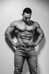 Naklejka premium A young athlete bodybuilder poses in the studio topless in jeans near the wall. Black and white.