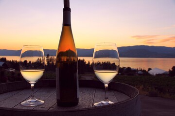 Dark sunset - romantic scene with the wine glasses and bottle of white wine on the wooden barrel by Okanagan Lake in Kelowna, BC - obrazy, fototapety, plakaty