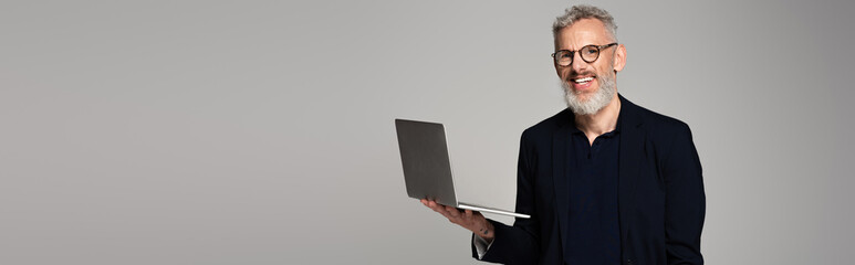 cheerful middle aged man in glasses holding laptop isolated on grey, banner - Powered by Adobe