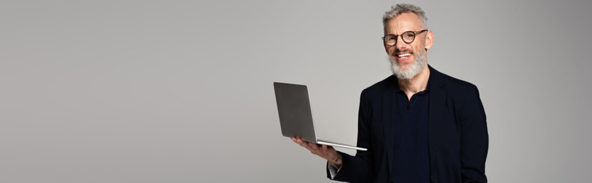 Cheerful Middle Aged Man In Glasses Holding Laptop Isolated On Grey, Banner