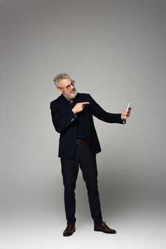 full length of businessman with grey hair pointing with finger while taking selfie on grey