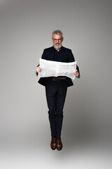 full length of middle aged businessman in glasses and suit reading economic news while levitating on grey