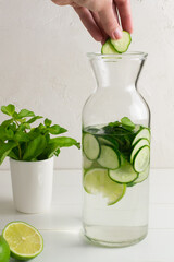 A hand tosses a few slices of cucumber into a jug of fresh water infused with cucumbers and mint sprigs, a detox drink. Health care, fitness, healthy food concept.