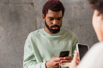 African american man using cellphone near cup of coffee and blurred girlfriend in cafe