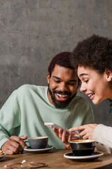 Positive african american couple using smartphone near cups of coffee in cafe