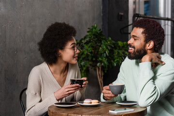 Smiling african american couple with coffee talking near macaroons in cafe