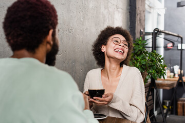 Positive african american woman with coffee sitting near blurred boyfriend in cafe