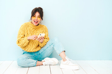 Young beautiful smiling female in trendy summer yellow hipster sweater and jeans.Sexy carefree woman sitting near blue wall in studio.Positive model having fun. Looking at smartphone screen, using app