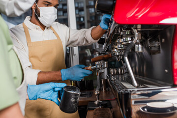 African american barista in medical mask making coffee on coffee machine near blurred colleague