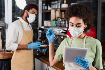 African american barista in medical mask and latex gloves using digital tablet and pointing at blurred colleague