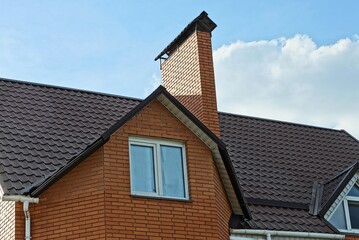 red brick attic with a white window and one large chimney on a brown tiled roof against a background of blue sky and clouds