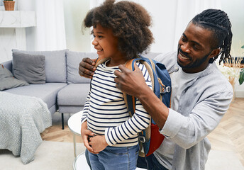 Happy African American daddy helps daughter getting ready for school and puts a school bag on her...