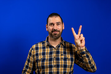 Handsome bearded man in a yellow plaid shirt making the victory gesture while looking at camera smiling isolated on blue studio background