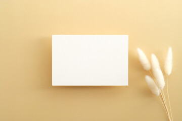 White invitation card mockup with dried flowers on beige background. Flat lay, top view, copy space