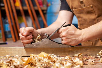 Carpenter in an apron planing a wooden board with a planer, workplace.