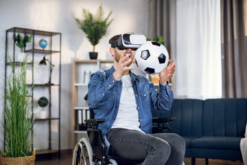 Cheerful young guy sitting in wheelchair using virtual headset for playing football game at home....